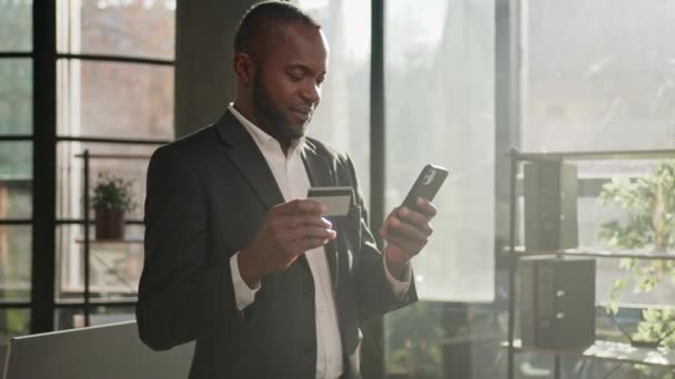 成熟した民族のビジネスマンアフリカ系アメリカ人40代の男性オフィスに立つ携帯電話のクレジットプラスチックカードを使用ウェブストアでオンライン決済を購入支払い銀行はコード番号を入力簡単にお金を転送 — ストック動画