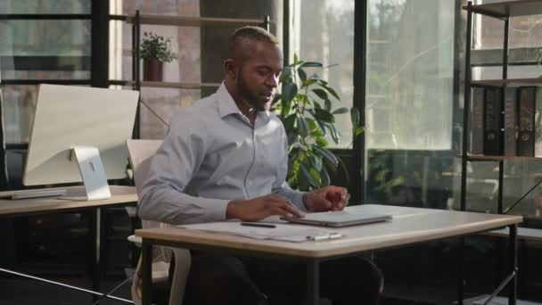 満足のいくビジネスマンアフリカ系アメリカ人成熟した中年45年代男性仕上げコンピュータプロジェクト近いラップトップ作業日オフリーンバック上の椅子に手を置きます後ろの頭は 太陽の暖かいオフィスでリラックス休憩を取ります — ストック動画