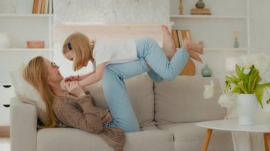 Happy Caucasian mommy playing with cute adorable toddler child daughter mother holding lifting funny little kid girl up having fun fly at mum knees gymnastics exercise airplane game on sofa at home