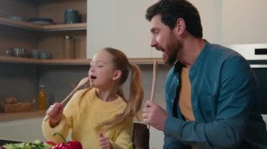 Kafkas sakallı komik bir baba ve mutfakta şarkı söyleyen sevimli küçük bir kız çocuğu komik bir aile babası ve elinde kaşıkla çocuk babası mikrofon şakıyarak yemek pişirmenin tadını çıkarın.