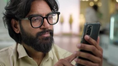 Indian male bearded man in glasses professional hold modern smartphone texting message in office indoors. Arabian businessman using mobile app browsing phone business chatting scrolling web online