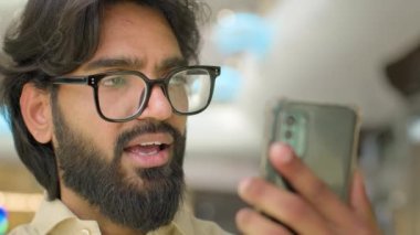 30s Indian bearded guy talk video call conversation on smart phone. Modern wireless technology usage. Arabian man in eyeglasses businessman talking to mobile web camera remote communication conference
