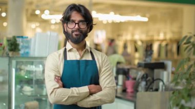 Portrait small business owner male bearded Indian Arabian man in eyeglasses standing in cafeteria restaurant posing arms crossed. Cafe waiter worker guy coffee service looking at camera crossing hands