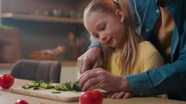 Happy Caucasian bearded father dad teach cute small kid girl child hold knife cut fresh cucumber vegetable for salad in kitchen help little daughter learn cook prepare healthy food cooking at home