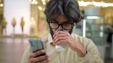 Male Indian bearded millennial professional hold modern smartphone drinking coffee in cafe. Arabian businessman in glasses man use mobile app for business look at phone drink tea at break in cafeteria