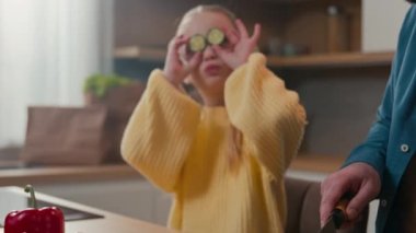 Caucasian adult father cut vegetables for salad with knife little daughter child girl cover eyes holding cucumber slice like glasses playing fooling together while cooking vegetarian food in kitchen