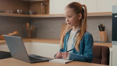 Distance learning at home kitchen. Adolescent schoolgirl Caucasian little child school girl studying online using laptop writing notes in copybook listen distant Internet teacher video lesson class