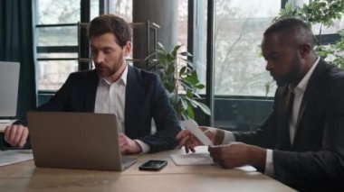 Diverse professional businessmen Caucasian man and African American businessman multiracial partners men with papers documents and laptop talking in office discuss online project at business meeting