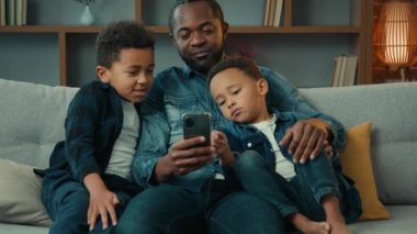 Happy African American family adult father with cute school kids children boys sons relax on sofa using funny smartphone apps talking use technology together looking at mobile phone play game at home