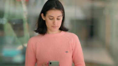 Serious Caucasian multiracial businesswoman multiethnic woman lady female girl indoors in office shopping mall scrolling phone texting chatting with mobile smartphone online browsing looking at camera