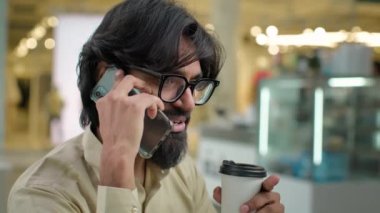 Indian bearded professional in glasses Arabian businessman talking business call talk on mobile phone in cafe drinking coffee tea work break pause man in eyeglasses speak smartphone drink indoors