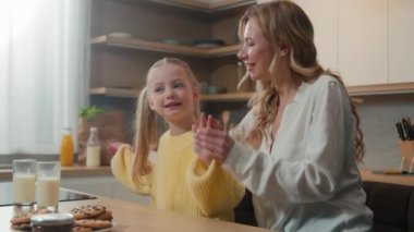 Caucasian child girl sitting with mother at dining table in kitchen having fun together, mom holding kid daughter by hands dancing to music moving with arms dance hugging cuddling, family breakfast