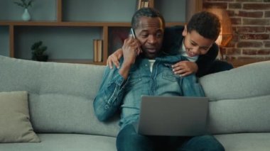 Adult African American father with laptop working at home distant job talking mobile phone sit on couch at living room little child son hug embrace daddy man from back children kids boys hugging dad