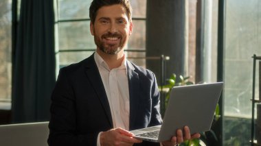 Caucasian businessman typing on laptop checking email standing in office morning in sunbeams sunrise man successful entrepreneur professional manager leader corporate executive smiling looking camera