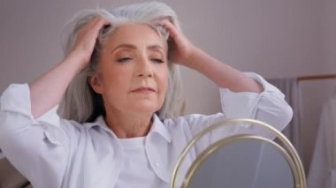 Kapatın yaşlı olgun kadın büyükanne modelinin tadını çıkarın. 60 'lı yaşlarda, beyaz kadın iyileşiyor. Uzun saçlar sağlıklı gri saç bakımına dokunuyor ve aynaya bakıyor.