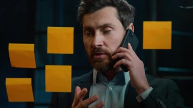 Caucasian businessman marketing assistant middle-aged man multitask talk mobile phone discuss project at office read sticky notes on glass manage business tasks goals analyzing sales strategy research
