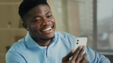İş videosu, gülümseyen Afro-Amerikan iş adamı cep telefonuyla konuşuyor. İnternetteki konferans uygulamasını akıllı telefondan kullanın. Milenyum etnik erkek girişimci ofis çalışanı içeride sohbet ediyor.