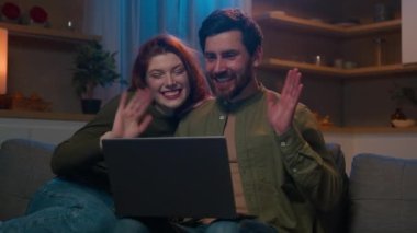 Happy young caucasian family couple sit on couch at home chatting with friends or parents via video call using laptop man and woman talk at online conference enjoy virtual communication waving hello