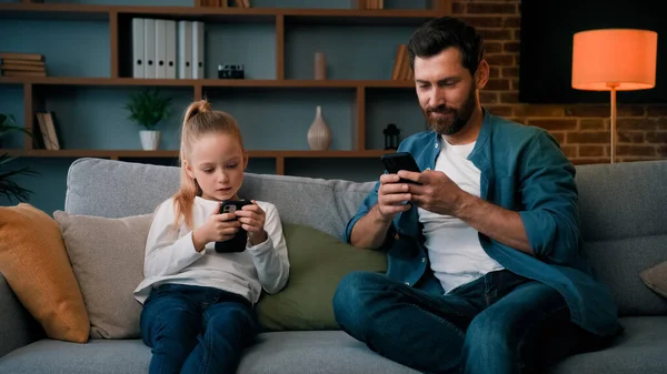 Caucasian single daddy and child kid preschooler girl sit on sofa look at mobile phone screen chatting online use home wifi smartphone apps young family internet addicted modern gadgets tech overuse