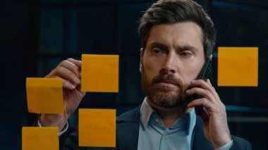 Caucasian businessman marketing manager middle-aged 40s man talk mobile phone multitask working in night office write sticky notes on glass wall data market analysis research business planning project