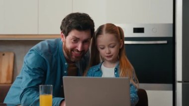Adult dad explain teaching small kid girl help with using laptop at home kitchen. Daughter learning typing surfing browsing internet with parental control using modern computer together with father