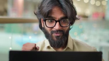 Excited Indian bearded business man in glasses reading great news on laptop celebrate online winning victory. Arabian businessman executive winner win with Internet job opportunity bonus achievement