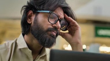 Close up worried stressed Indian bearded man in glasses suffer from head high pressure headache painful head thinking problem solution with laptop at workplace. Exhausted tired Arabian businessman
