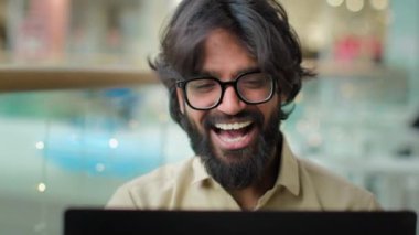 Happy executive Indian bearded entrepreneur man in glasses celebrate win with laptop achievement talk video call shout with success. Arabian businessman winning business goal achieve online victory