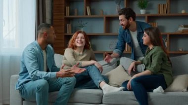 Four ethnic friends fellows companions sit on home couch talking discuss plans chatting friendly talk indoors gathered multiethnic men and women converse joking laughing sharing news real friendship