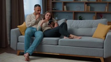 Millennial 30 'ların çifti cep telefonlarını kullanarak koltukta dinleniyor uzaktan satın alma hizmetleri sunuyor çeşitli Afro-Amerikan erkek ve beyaz kadın kocaları internette geziniyor.