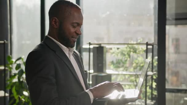 太陽の光でラップトップと一緒にコンピュータのブラウジングウェブサイトに入力する成熟した男が立っています 中高年アフリカ系アメリカ人実業家男性エグゼクティブサーチ現代オフィスで働くデジタルデータ — ストック動画