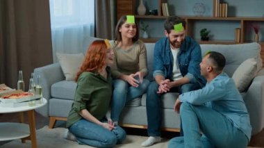 Four diverse friends play guessing game with sticky notes on head creative multiracial women men in living room playing intellectual charades asking questions Who am I talking laughing funny playtime