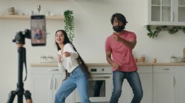 Interracial couple recording trendy dance move for social media ethnic arabian hispanic friends bloggers make video footage on mobile phone camera tripod at kitchen two funny dancers dancing to music