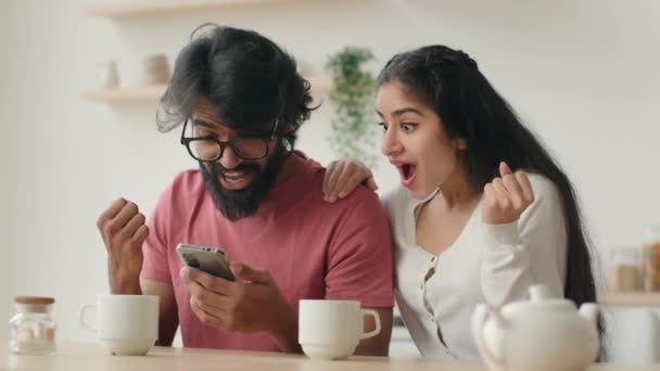 多人種カップルインド人男性と女性は スマートフォンの幸せな民族の配偶者でオンラインボーナスを獲得読んでニュースを楽しんで携帯電話アプリケーションを使用してキッチンに座って勝利を喜びます高い5を与えます — ストック動画