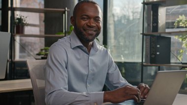 Professional middle-aged mature 40s ethnic African American man senior businessman office worker sit indoors search internet data on laptop pc device looking at camera friendly smiling positive smile