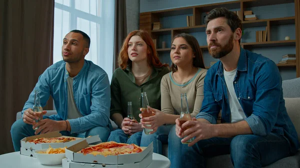Four friends watch television with beer and pizza at home diverse multiethnic women men sport fans supporters watching tv football match championship soccer game sad worried by team losing bad result