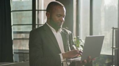 Afrikalı etnik iş adamı olgun orta yaşlı 40 'lı yaşlarda dizüstü bilgisayarlı güneş ışınları arka planda bilgisayar klavyesi erkek CEO' sunun internet sitesinde yazı yazarak duruyor.