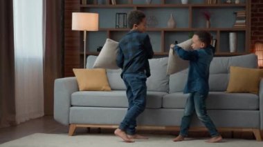 Two active playful little kids boys siblings fighting with pillows at living room. Ethnic brothers African American children friends at home having fun cheerful playing funny fight battle play game