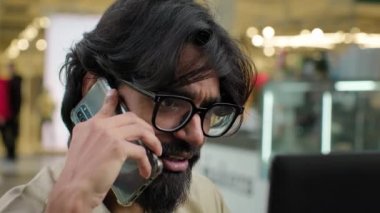 Smiling bearded Indian business man in glasses using laptop talking on cell phone sit at desk. Happy Arabian male businessman professional manager consulting client about online project answer call