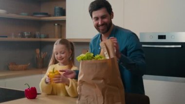 Little Caucasian child girl kid daughter help to adult man father unpack delivered vegetables in paper bag lettuce leaves fresh clean paprika tomato at home kitchen online supermarket shopping service