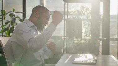 Emotional winner ethnic businessman African American adult mature 40s man celebrate victory win online bet on laptop business finance growth funny dancing at office desk yes gesture sunny background
