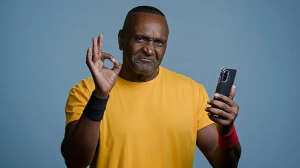 Mature african athletic man stands in studio gray background looking phone screen sportive male athlete trainer browsing mobile application for workout exercise sports showing approval sign gesture ok