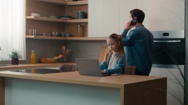 Caucasian man father dad talking mobile phone come in kitchen pouring juice in glasses while little girl schoolgirl pupil kid child daughter studying learning with laptop distant education from home