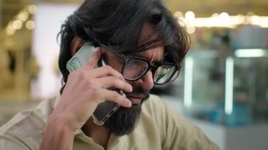 Stressed angry Indian bearded man in glasses talking mobile phone negative conversation awful news at call Arabian businessman entrepreneur quarrel conflict with client talk on smartphone screaming