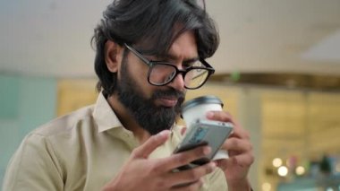Close up smiling Indian bearded millennial man in glasses drinking coffee scrolling browsing using smartphone in cafe. Arabian businessman drink tea use mobile phone app for business chatting online