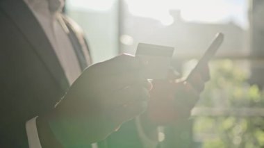 Close up male hands unrecognizable african american man hold mobile phone and plastic credit card send money web ecommerce use digital banking pay online. Unknown businessman stand in sun sunny office