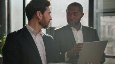 Caucasian man sales manager talking to African American male company client in office. Two diverse multiracial men employees workmates discuss business project software with laptop agree startup idea