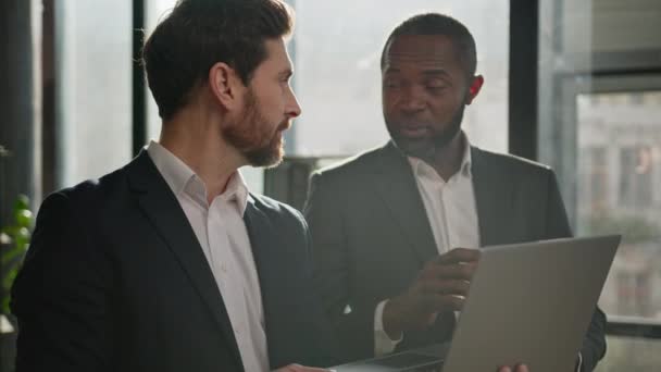 白人男性セールスマネージャーはアフリカ系アメリカ人男性の会社の顧客とオフィスで話しています 2人の多様な多人種の男性従業員がラップトップでビジネスプロジェクトソフトウェアを議論するスタートアップのアイデアに同意する — ストック動画