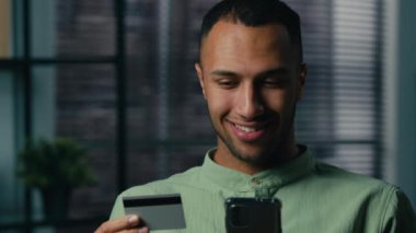 Gülümseyen Afro-Amerikan genç adam elinde kredi kartı tutarak cep telefonu bankacılığı uygulamasını kullanarak akıllı telefondan anında ödemeyle satın alan mutlu erkek müşteri alıcısı satın alıyor.