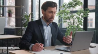 Concentrated businessman talking on video call using laptop in office man coach advisor consulting client writing notes having distance interview communicate at online conference with business partner